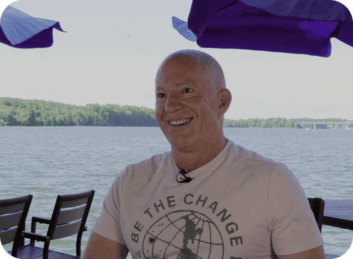 Amir Harpaz - the owner of The Mudpuppy, seated on a lakeside patio under blue umbrellas.