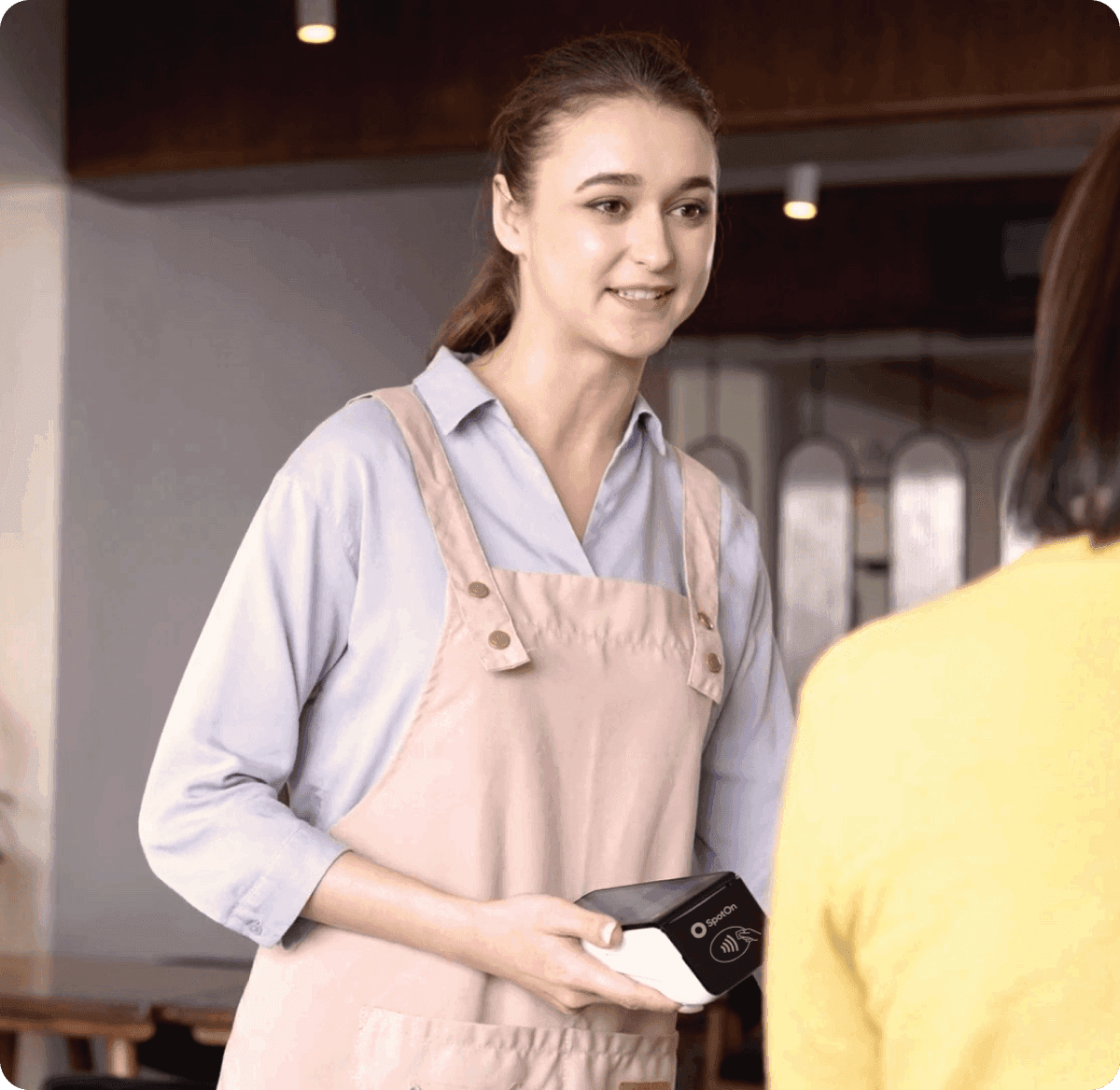 Server in light blue shirt and pink apron presenting SpotOn payment terminal to customer in restaurant setting