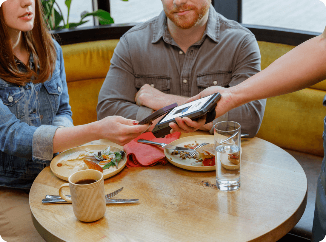 Customers at restaurant table with yellow banquette seating completing payment transaction, coffee cup and finished plates visible