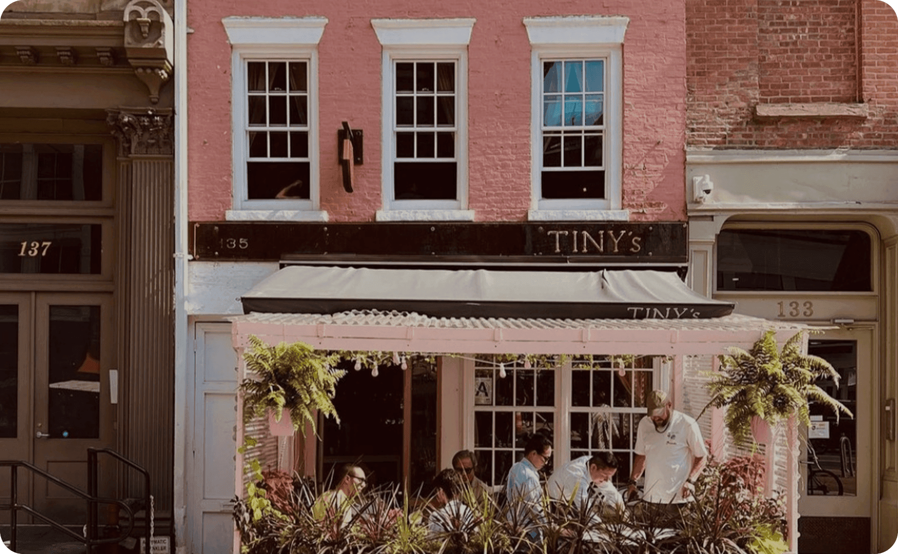 The pink townhouse exterior of Tiny’s & The Bar Upstairs, with guests dining on the covered patio