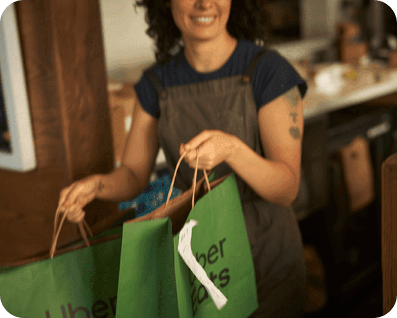 A smiling woman in an apron holds two green Uber Eats paper bags