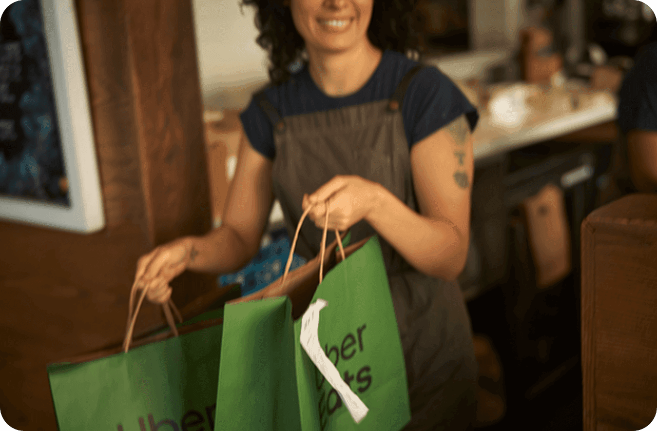 A smiling woman in an apron holds two green Uber Eats paper bags