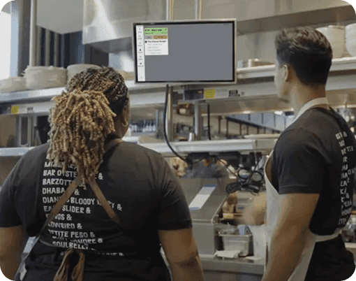 Two cooks look at a kitchen display system (KDS)