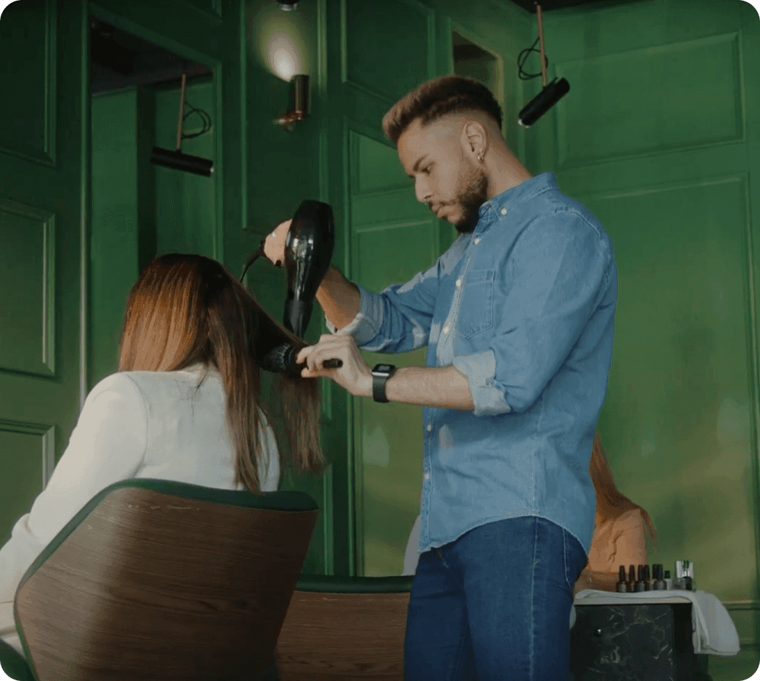 Professional hairstylist in a modern salon with green paneled walls styling a client's hair using a blow dryer