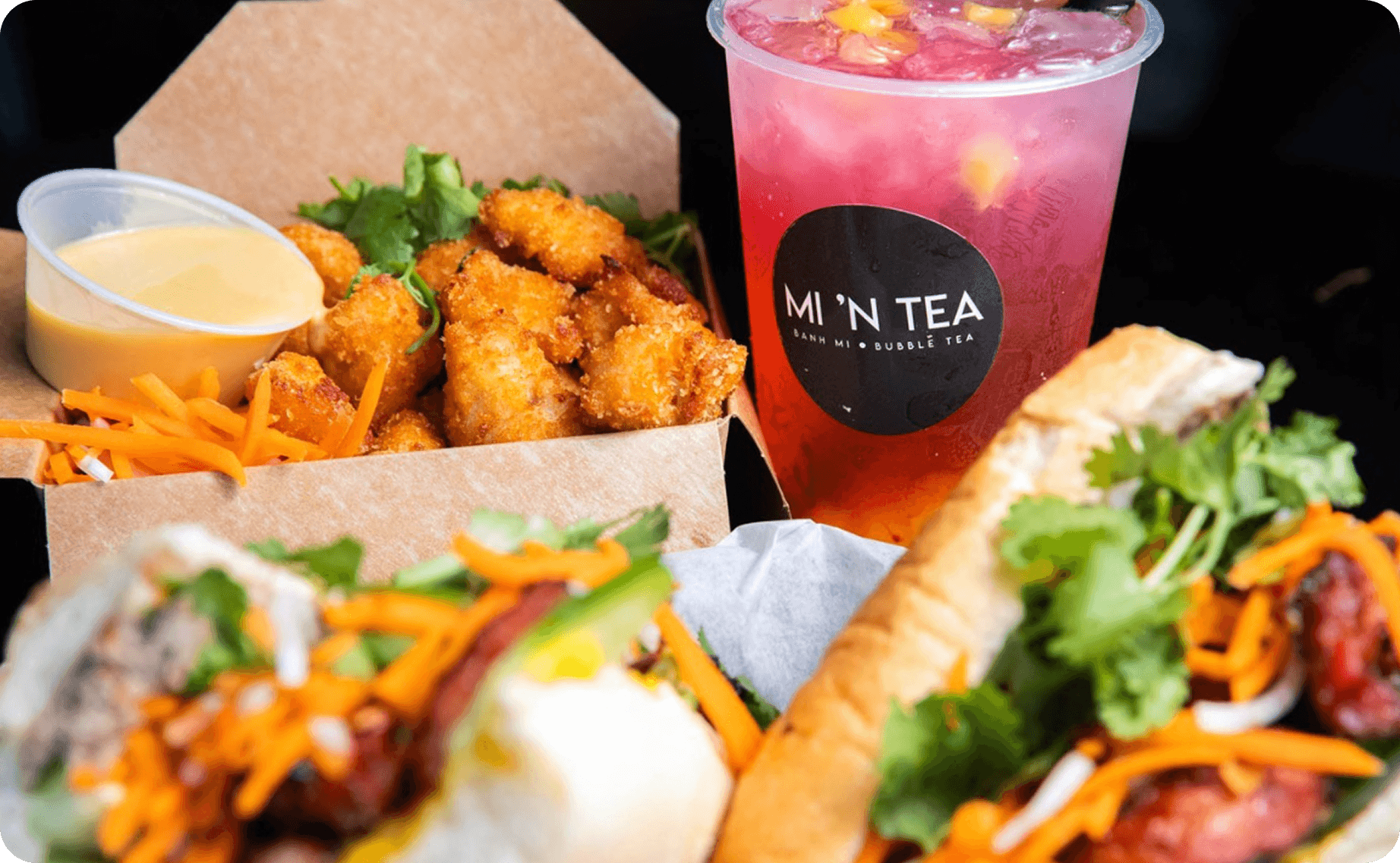 Bánh mì sandwiches, fried bites, and a pink bubble tea in a branded Mi ’N Tea cup