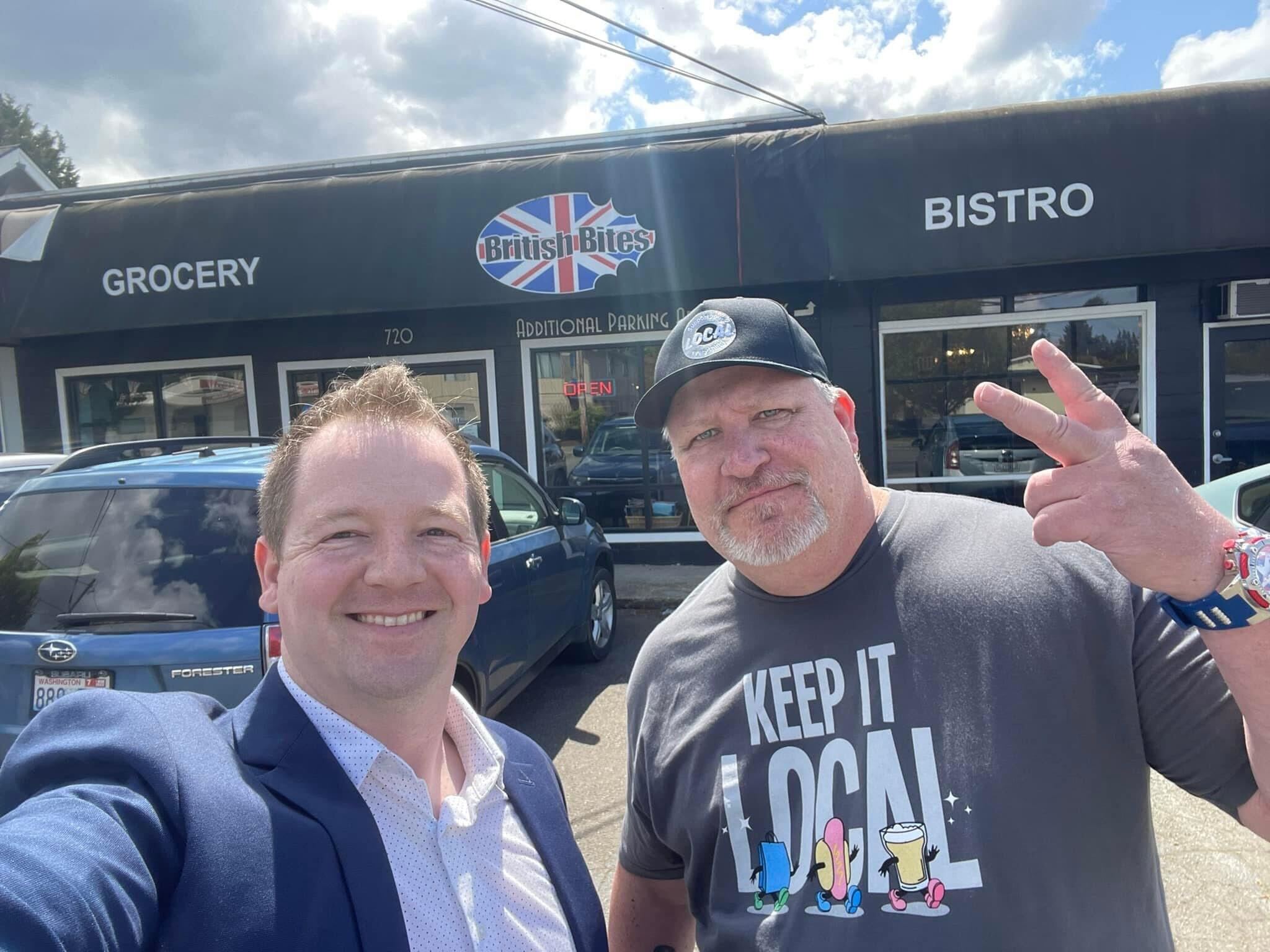 Two men pose in front of a restaurant, British Bites. The man on the right is wearing a hat and pointing two fingers up.