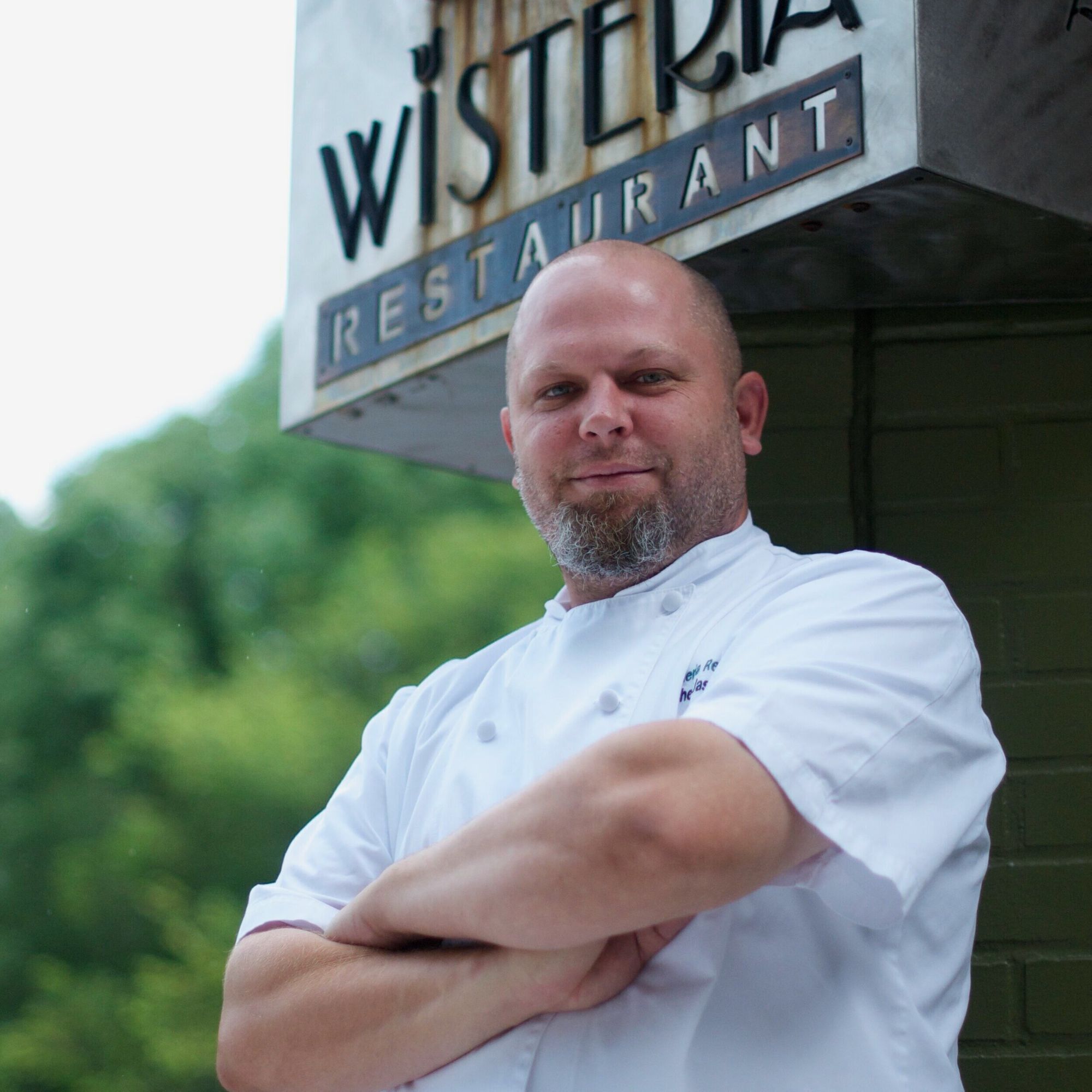 A man with a chef's coat smiles at the camera. His arms are crossed. The Wisteria Restaurant is in the background. 