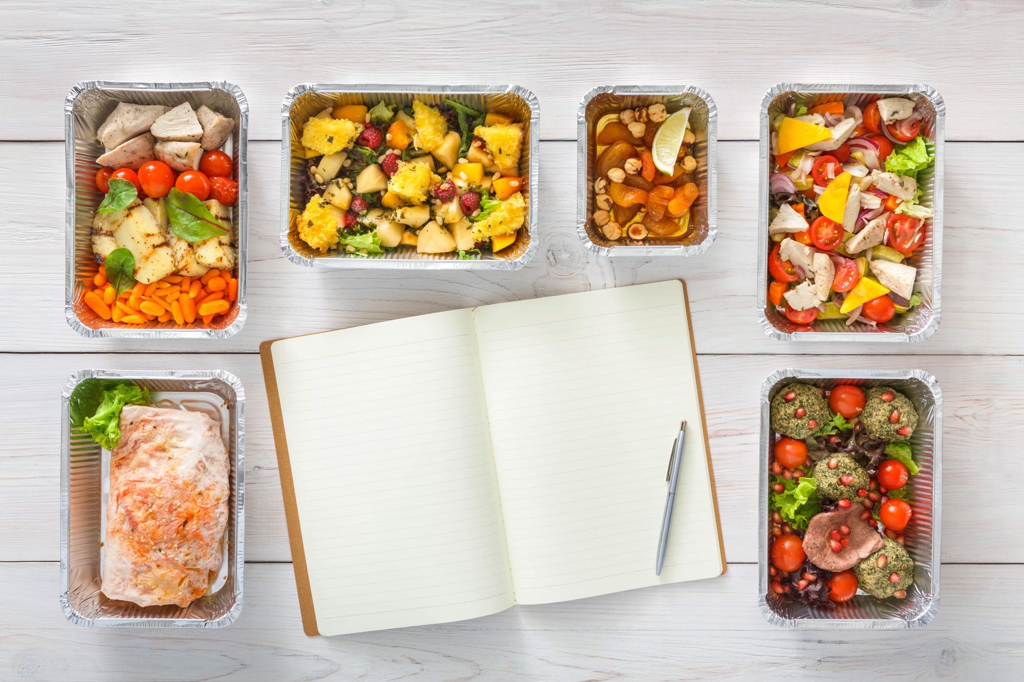 Six menu items in aluminum containers around a notebook and a pen.