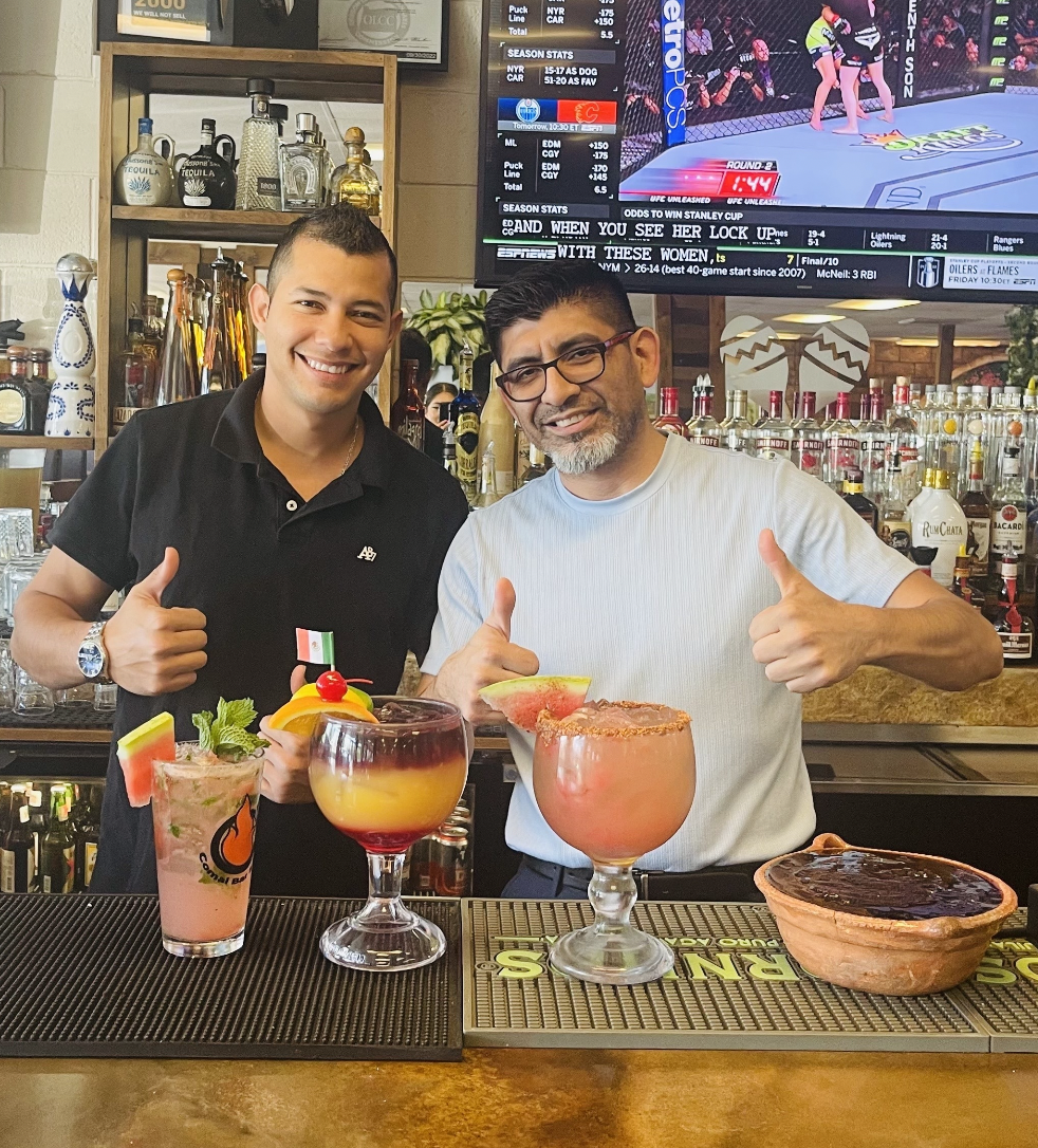 Two men stand in front of a bar and drinks, posing with two thumbs up.