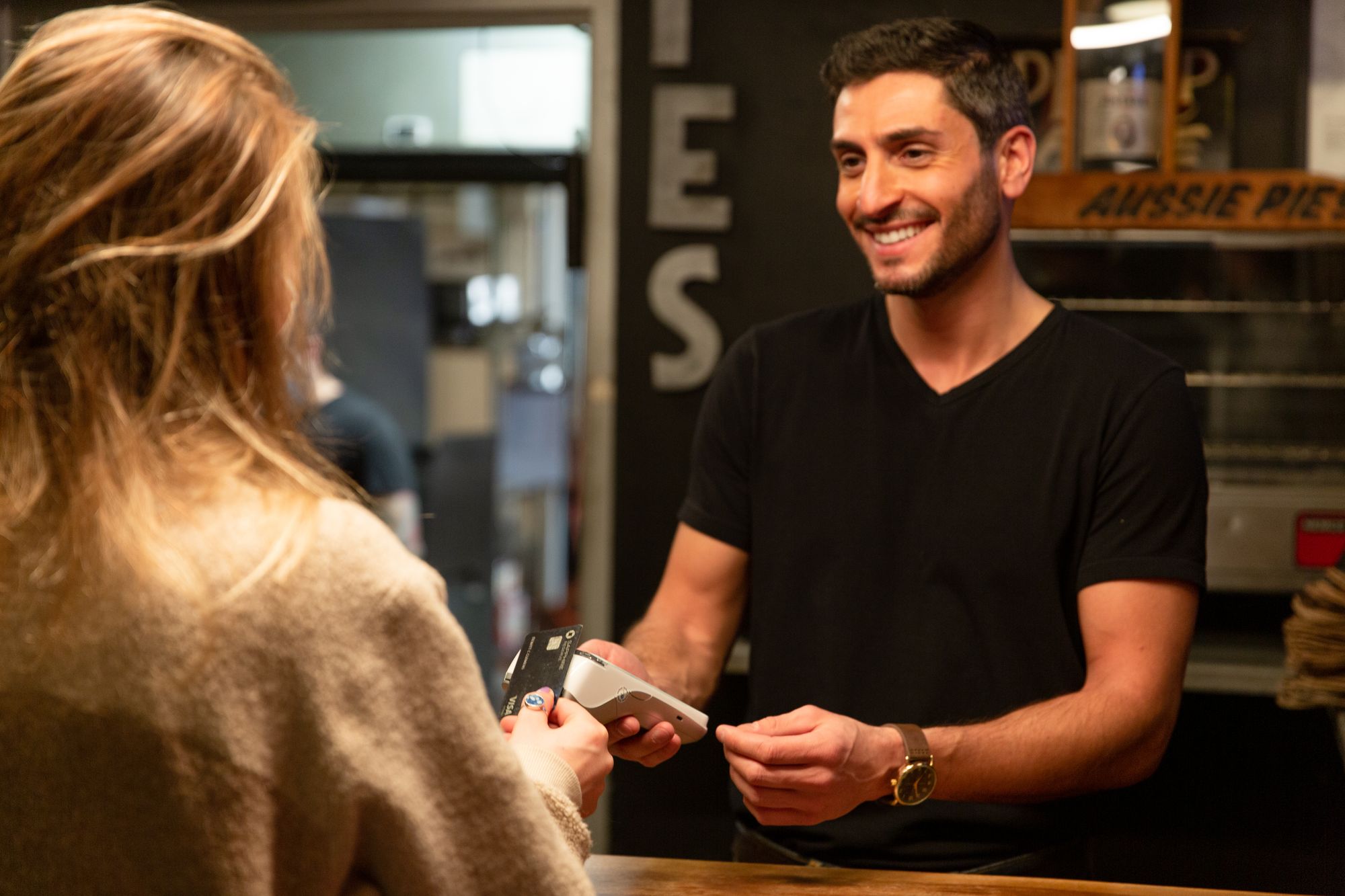 A restaurant waiter holds a handheld point-of-sale device. A customer uses her credit card to pay.