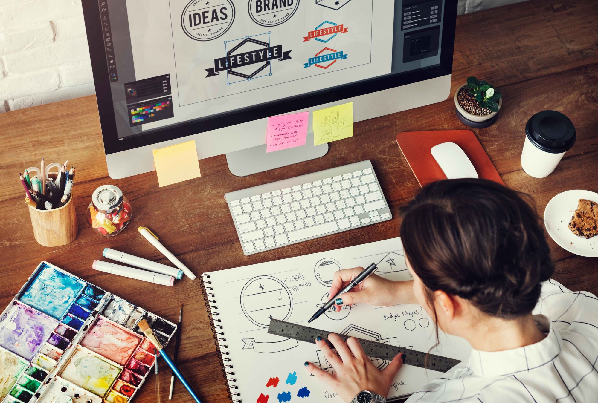 A graphic designer is sketching logo ideas at a desk in front of a computer.