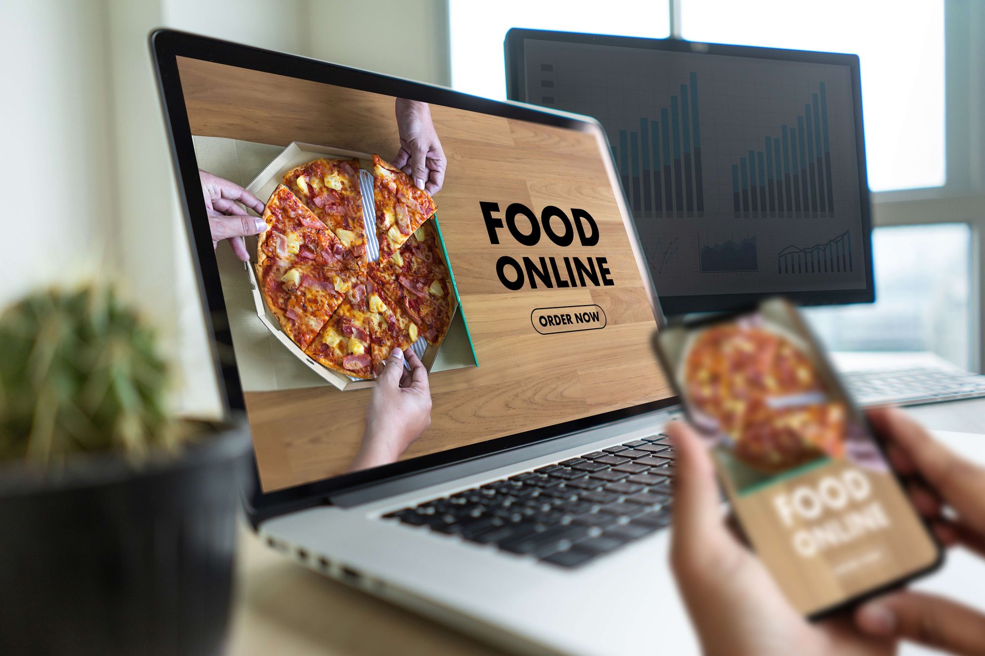 A hand holding a phone in front of a laptop with the words "Food Online" on the screens.