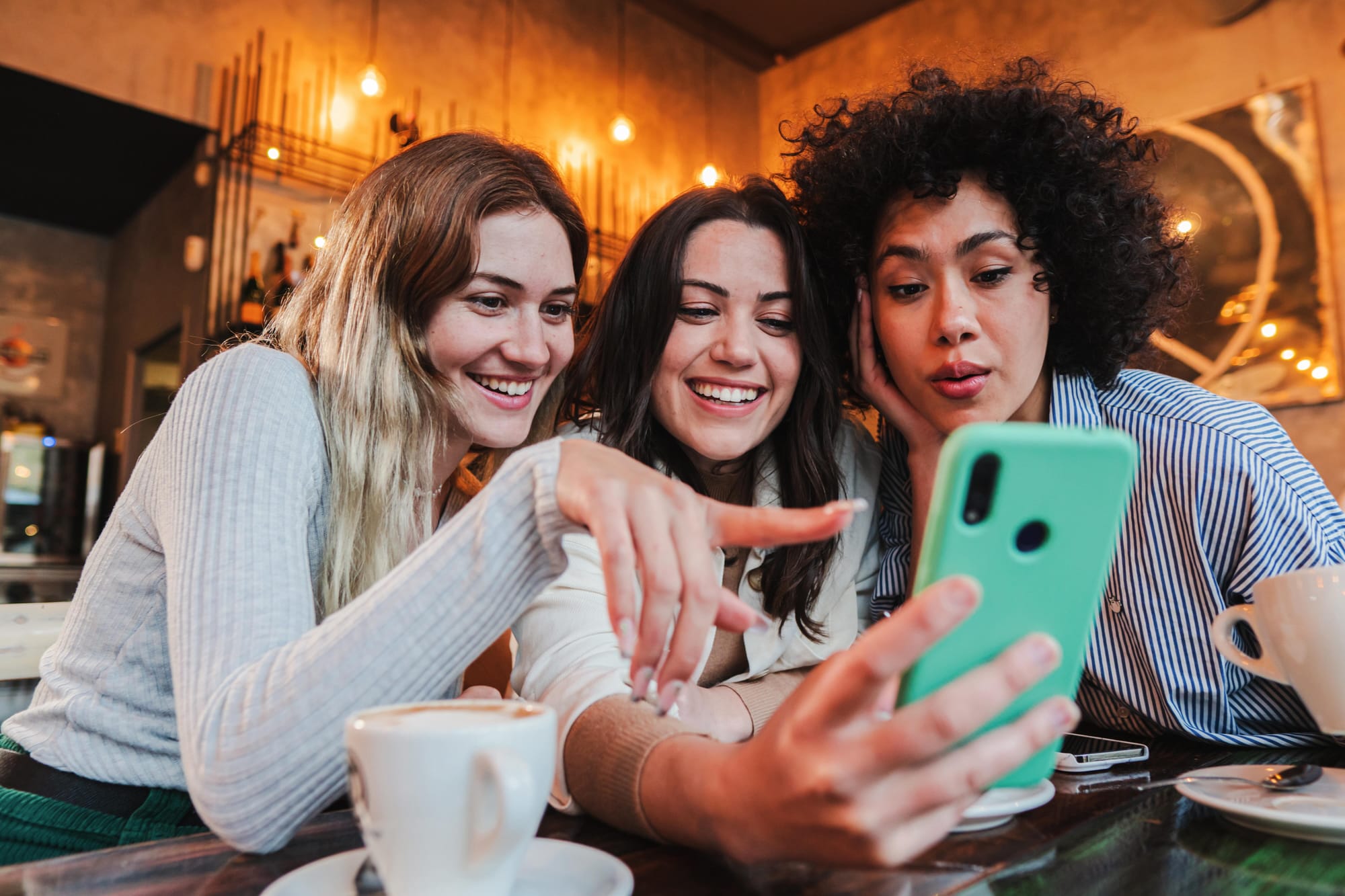 Three young women are smiling and looking at a phone in a restaurant.