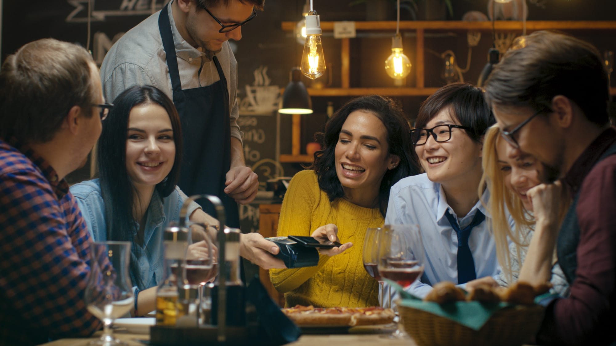 6 guests are at a table in a restaurant while a woman pays with her phone.