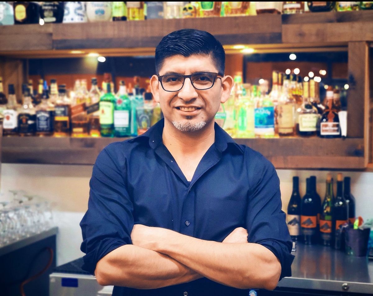 Man in glasses and small beard stands in front of a full bar with his arms crossed.