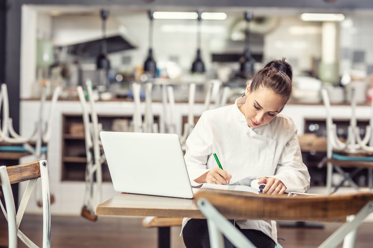 A chef sits at a table after hours in her restaurant and writes in a notebook with her laptop open. 