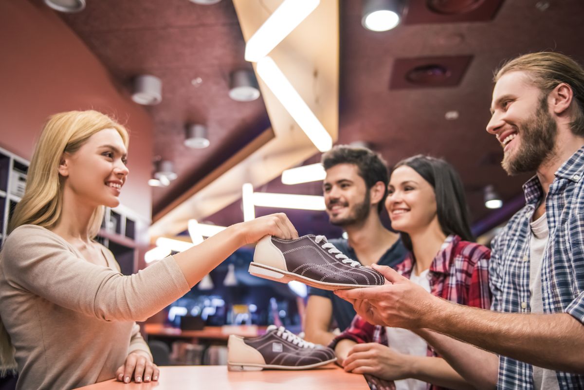 Woman in a bowling alley handing bowling shoes to a smiling customer.