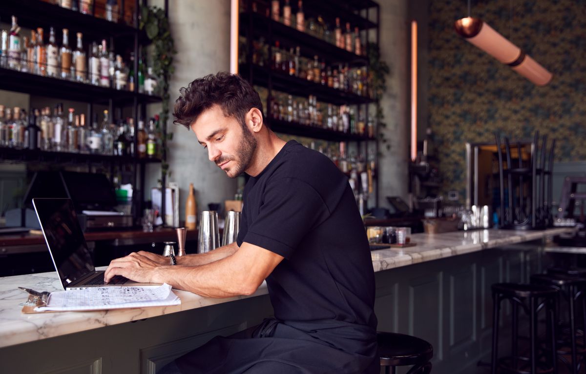 A man is sitting at the counter of a restaurant and examining a piece of paper as he types on a laptop.