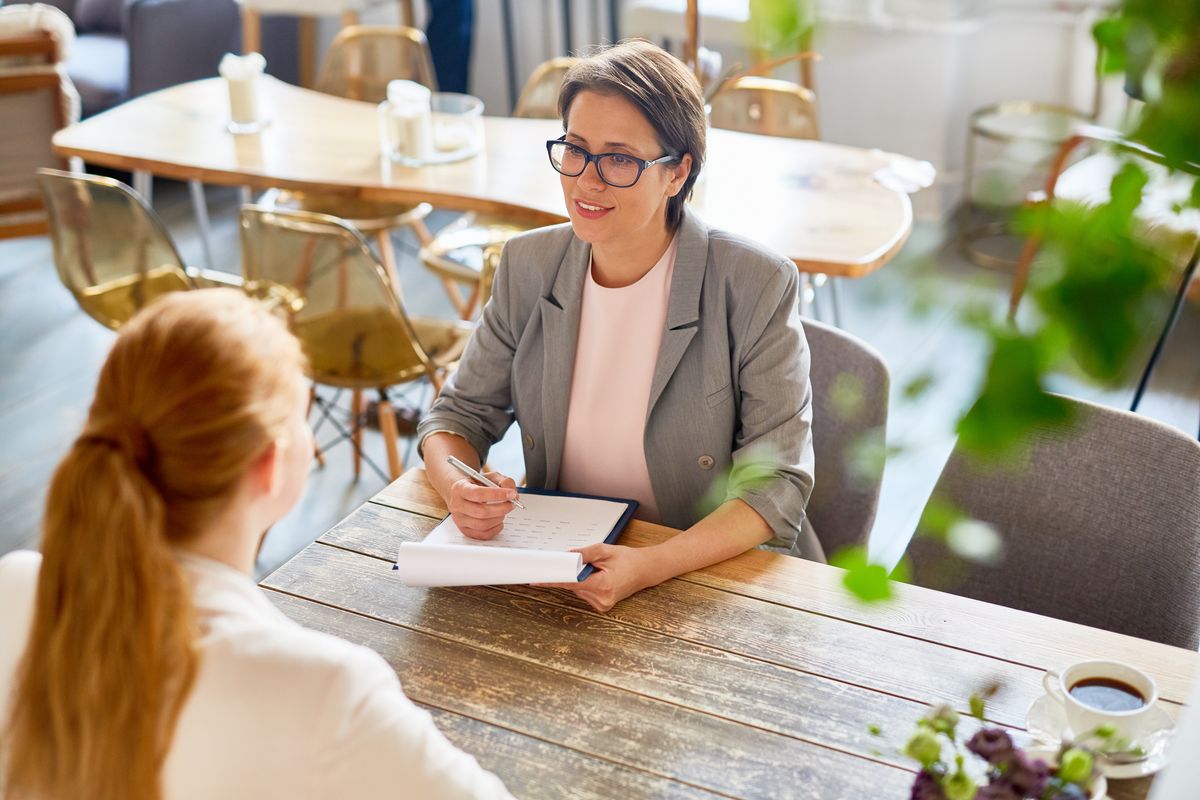 A manager in a new restaurant holds a paper and pen, interviewing a new restaurant staff member.