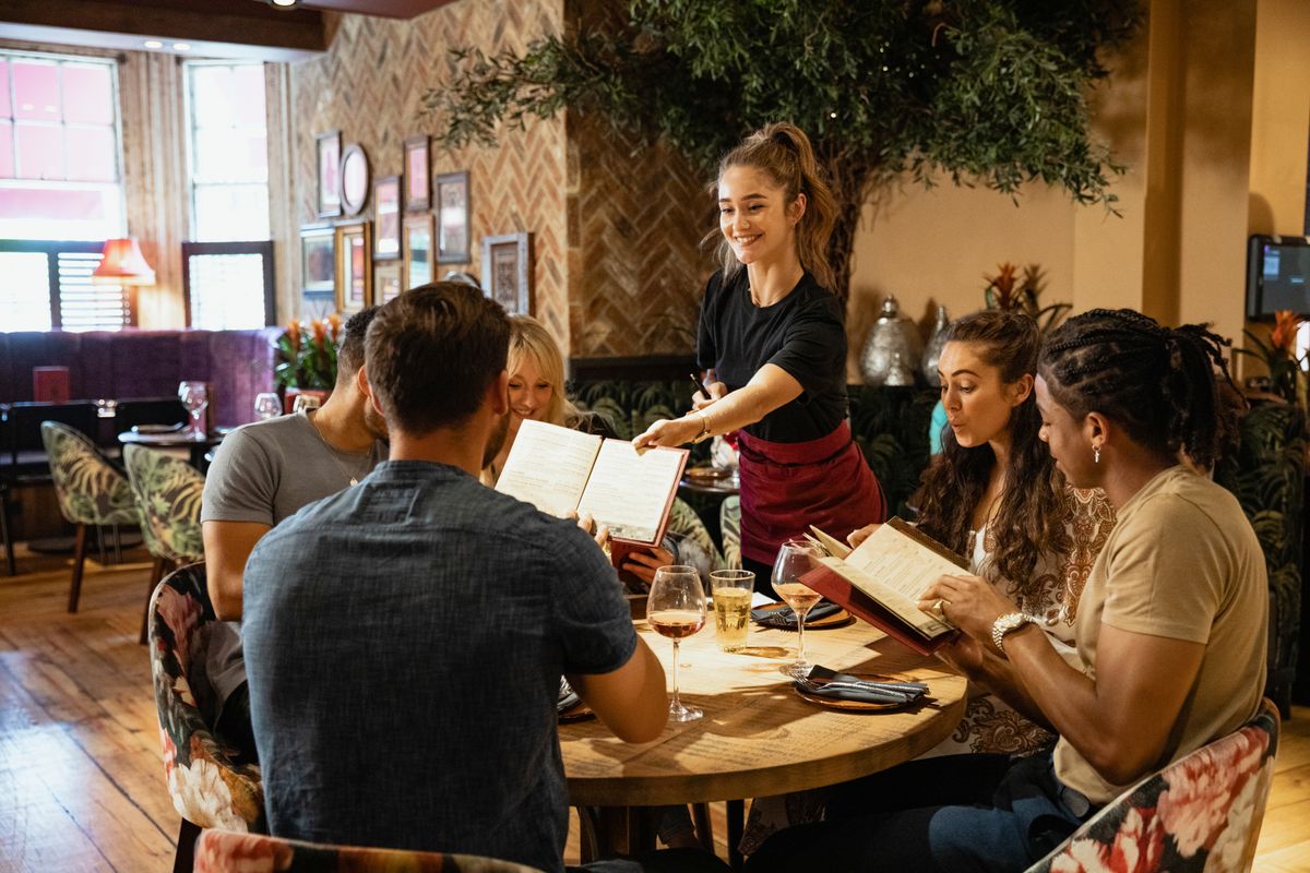 Server at table smiles and hands menu to guests seated at a restaurant, eager to order.