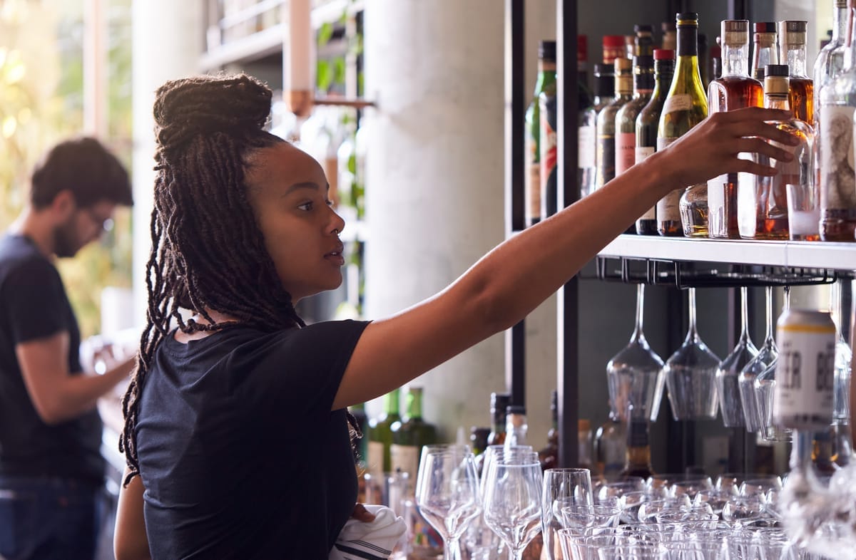 Woman barback reaches for alcohol from a shelf behind the bar.