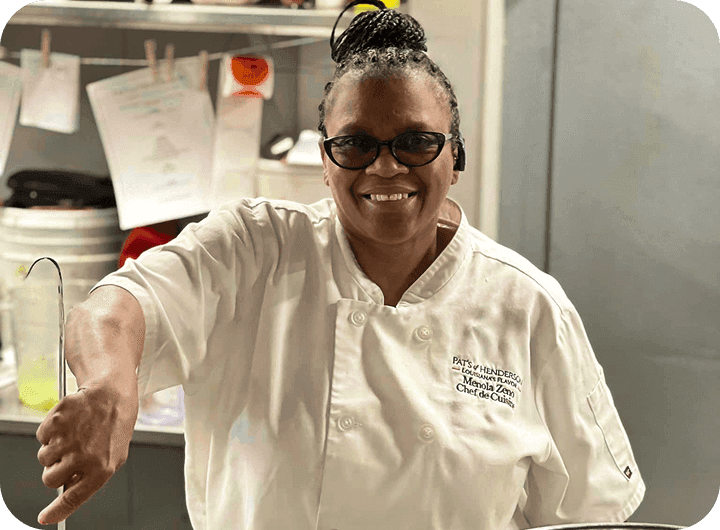 A smiling chef stirs a hot dish in a large pot in a commercial kitchen.