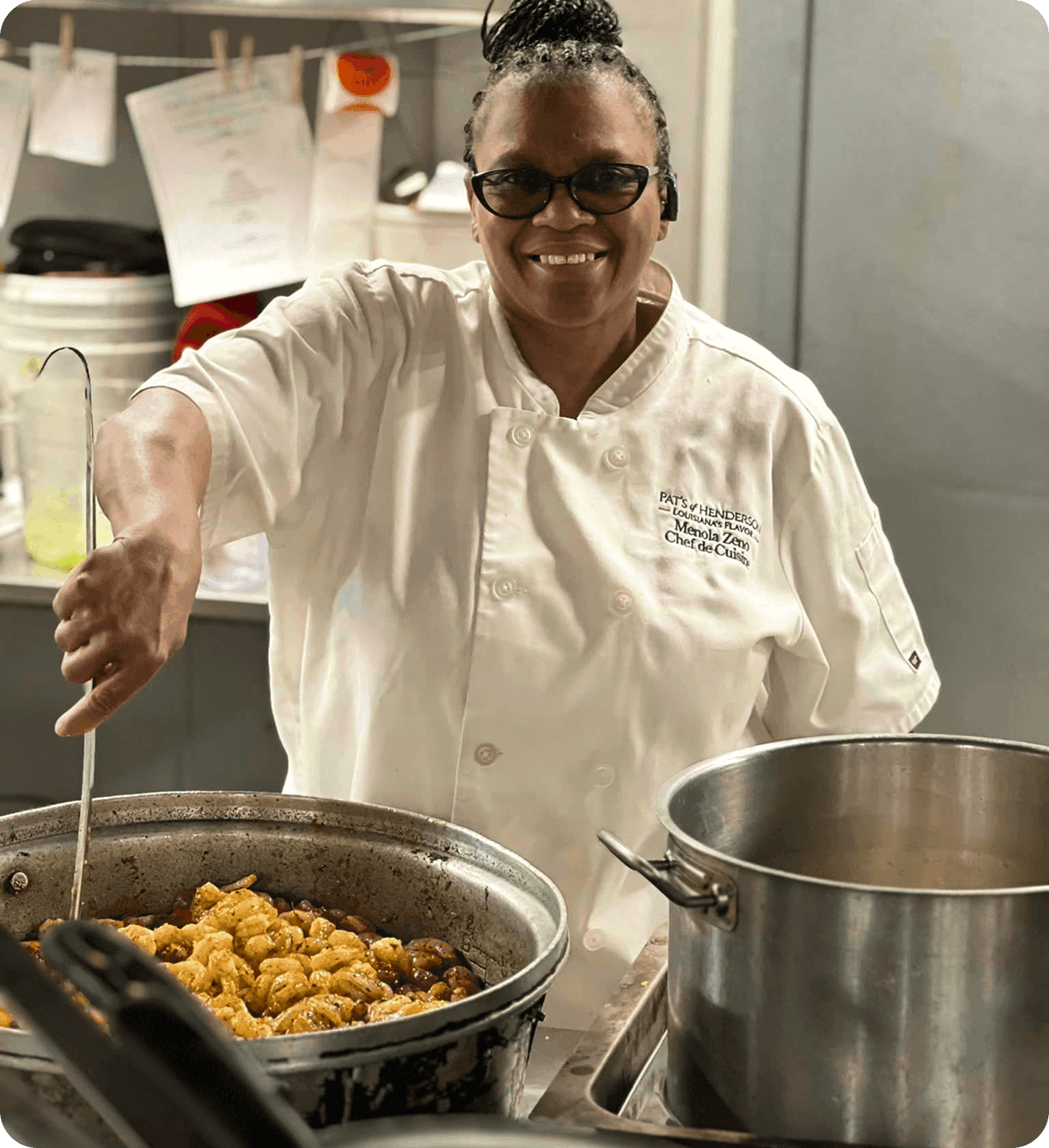 A smiling chef stirs a hot dish in a large pot in a commercial kitchen.