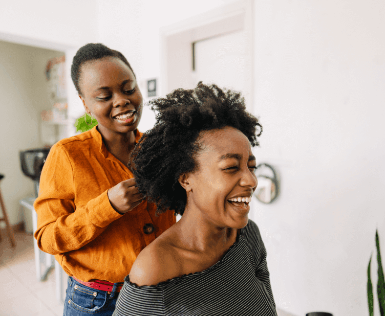 Two people sharing a genuine moment of joy during hair styling session, bright natural lighting and indoor plants visible in background