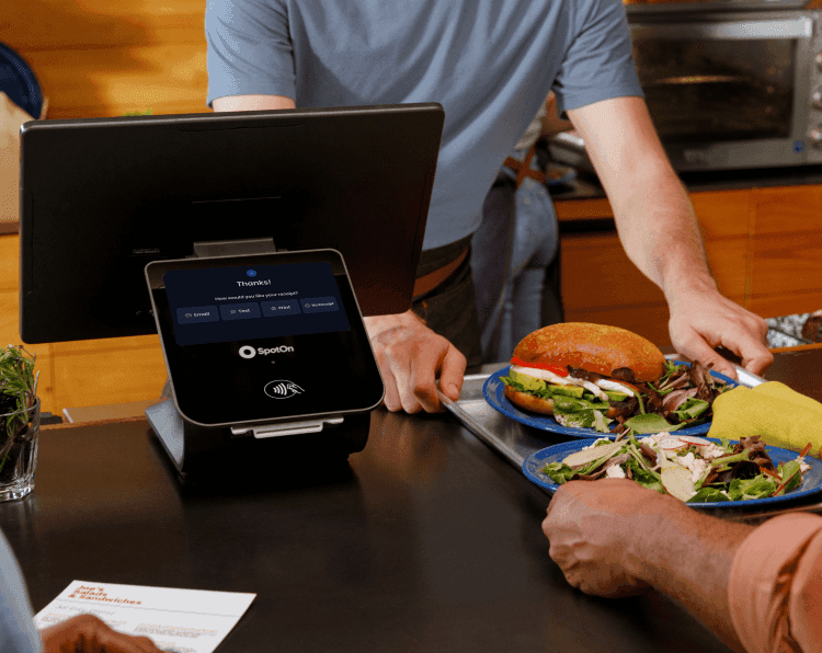 Restaurant staff demonstrating digital menu system on tablet POS to customers, with fresh salads and sandwiches displayed in counter case