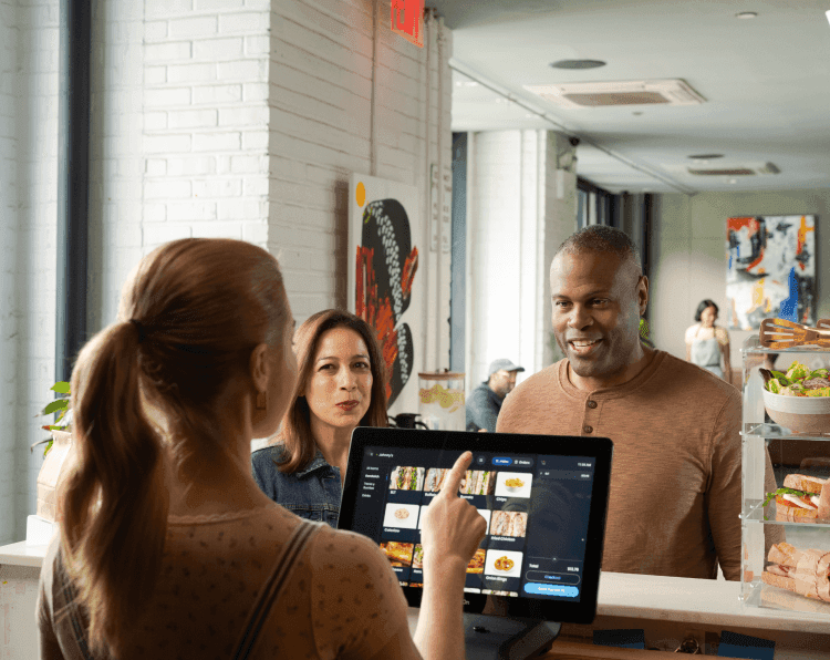 Restaurant staff demonstrating digital menu system on tablet POS to customers, with fresh salads and sandwiches displayed in counter case