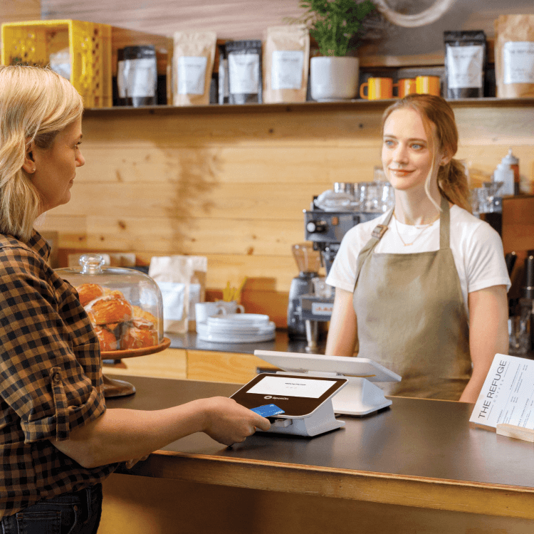 Cafe counter transaction between customer and barista, featuring SpotOn payment terminal, pastry display case, and shelves lined with coffee bags and orange mugs