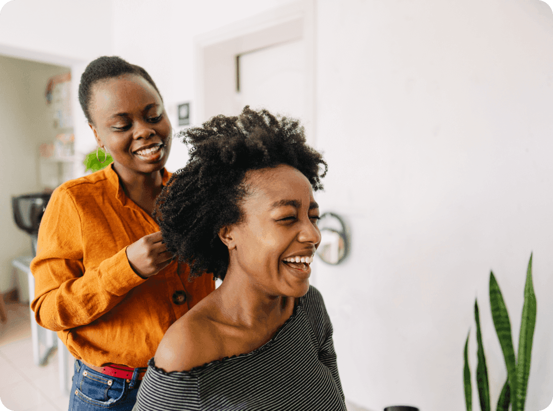 Two people sharing a genuine moment of joy during hair styling session, bright natural lighting and indoor plants visible in background
