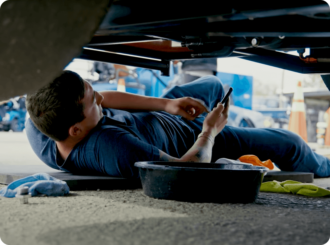 Auto mechanic checking mobile device while inspecting underside of vehicle, with maintenance tools and equipment visible