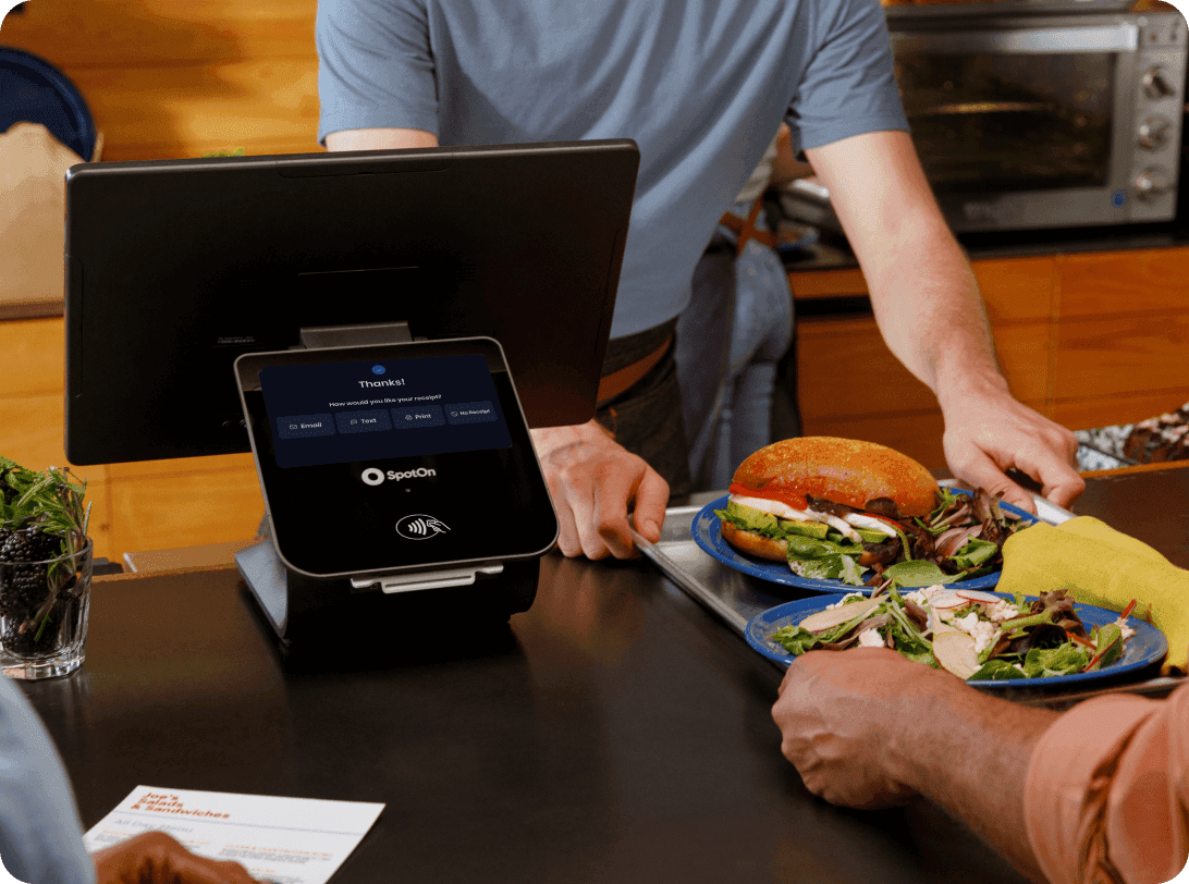 Restaurant staff demonstrating digital menu system on tablet POS to customers, with fresh salads and sandwiches displayed in counter case