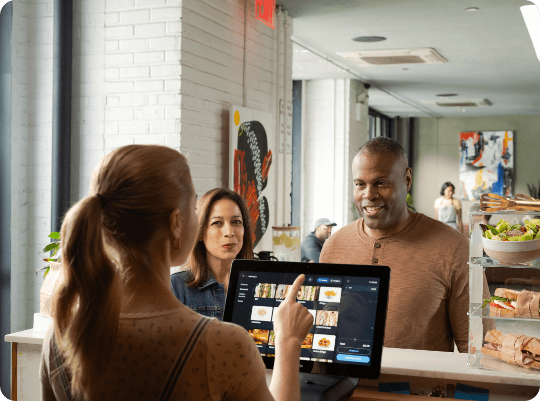 Restaurant staff demonstrating digital menu system on tablet POS to customers, with fresh salads and sandwiches displayed in counter case