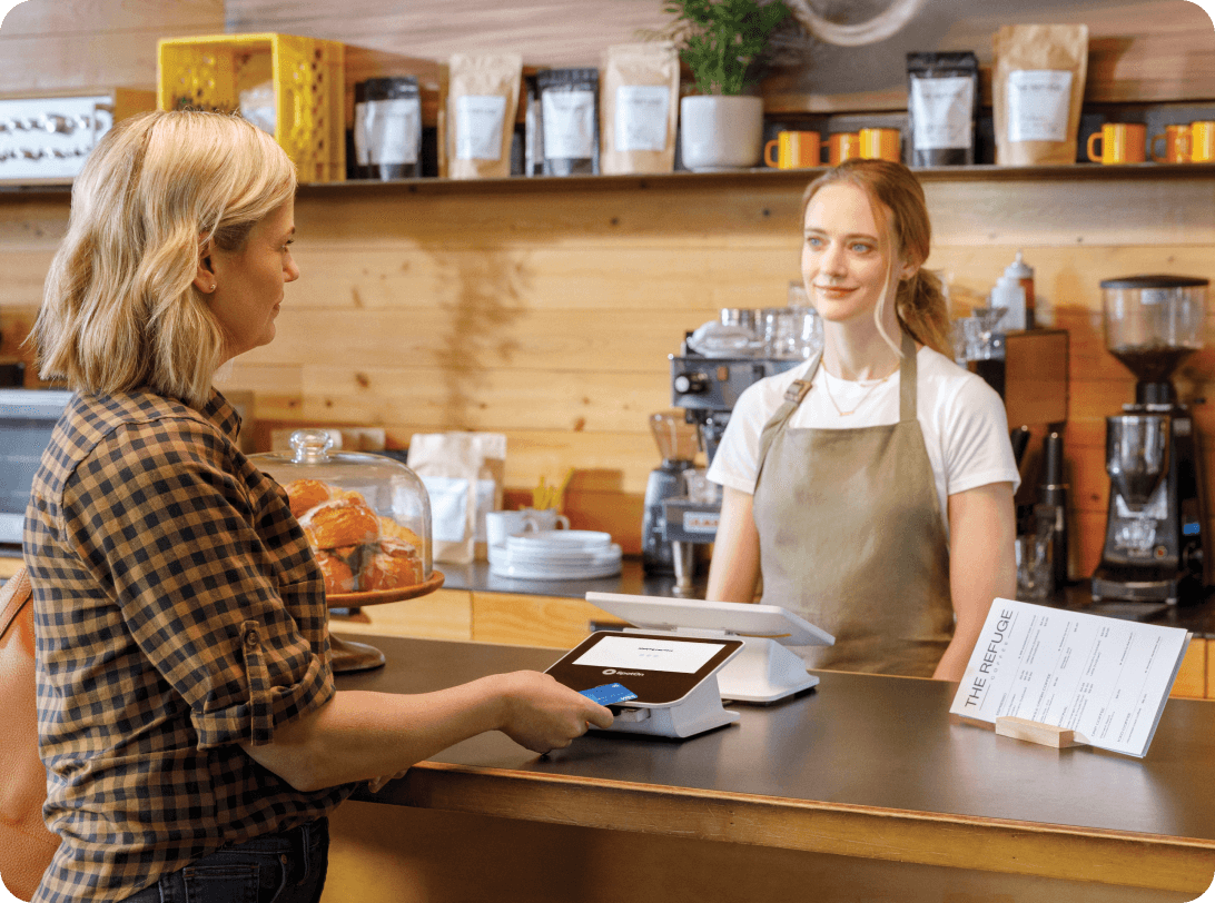 Cafe counter transaction between customer and barista, featuring SpotOn payment terminal, pastry display case, and shelves lined with coffee bags and orange mugs