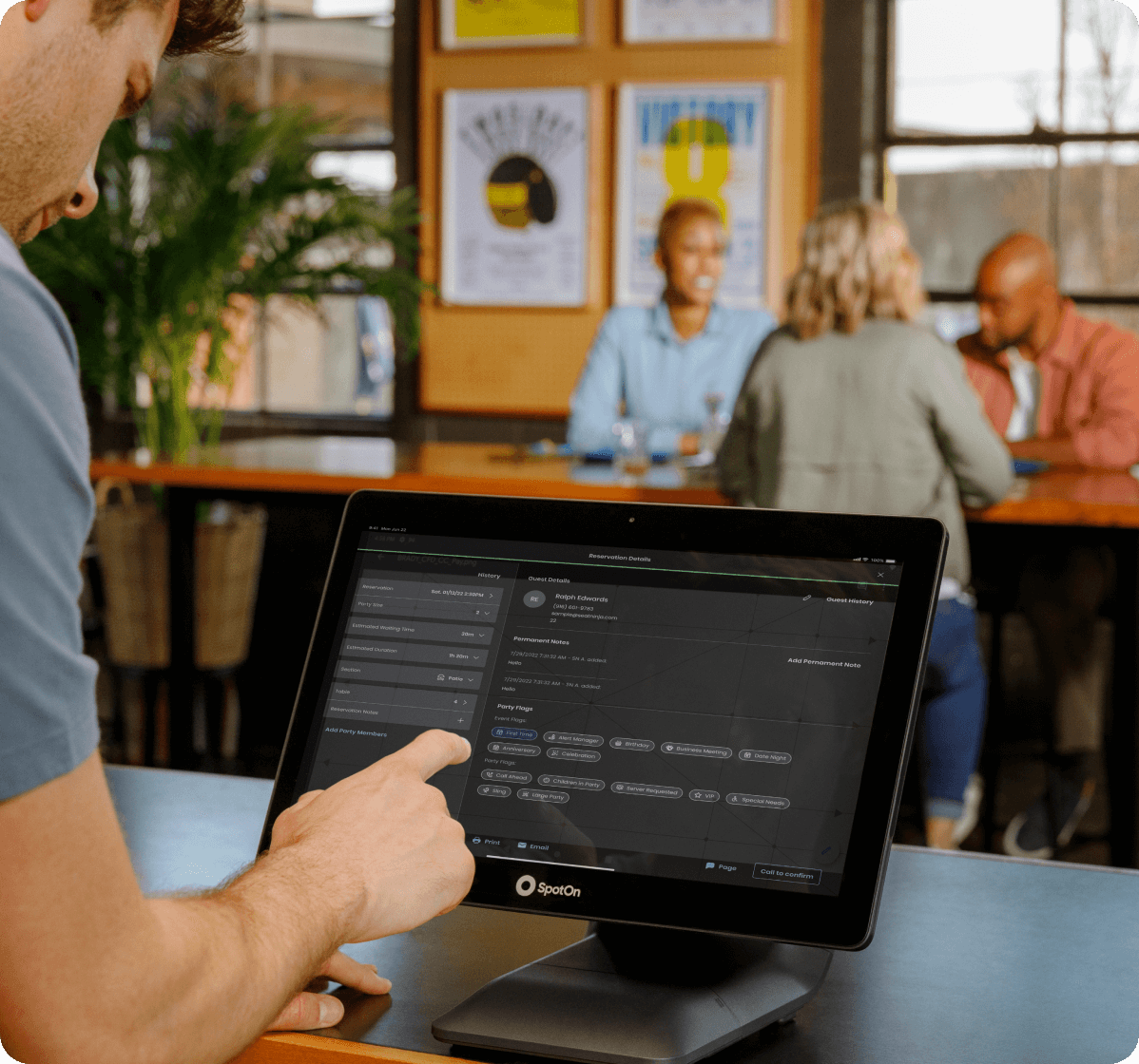 Restaurant staff member using SpotOn tablet POS system while customers dine in background, with dark mode interface visible on screen