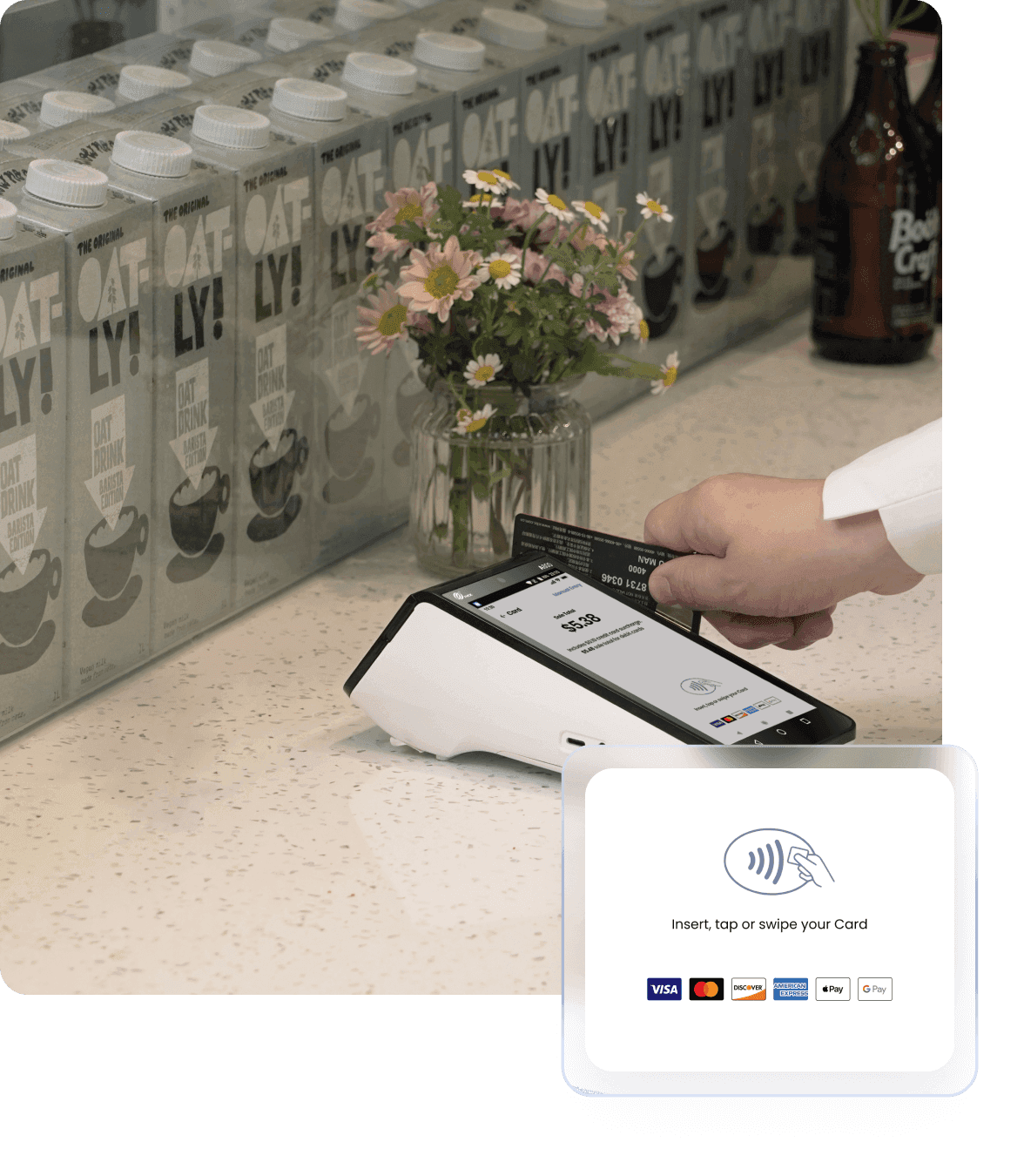 Payment terminal at cafe counter with rows of oat milk cartons in background, terminal displaying payment interface with card being processed