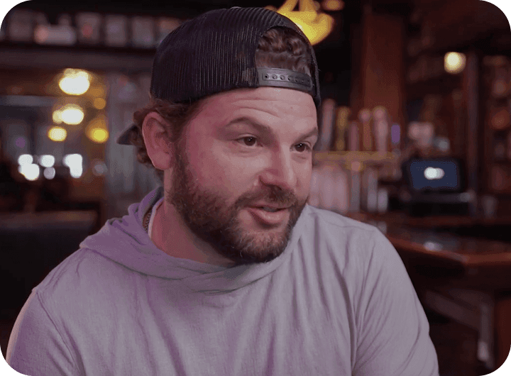 Man in a backward cap talking in a warmly lit bar.