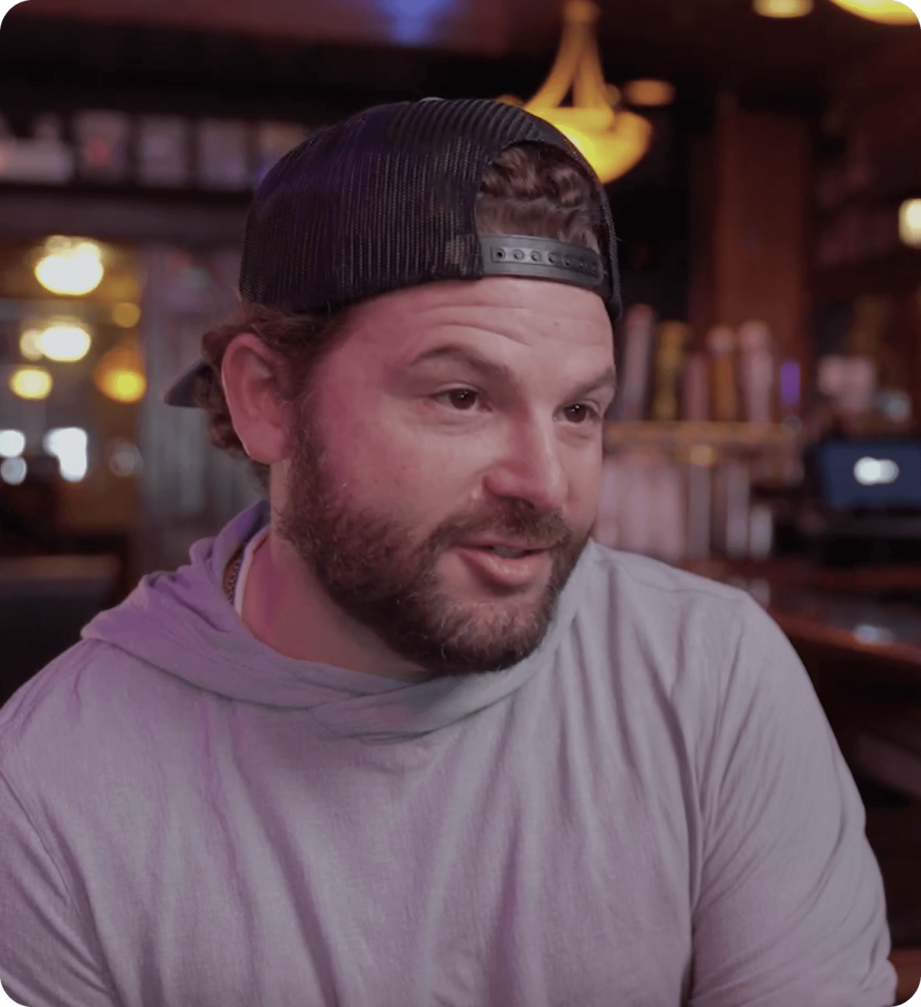 Man in a backward cap talking in a warmly lit bar.